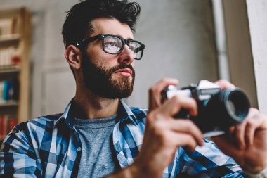 Düşünceli hipster adam giyim gözlük vizyon düzeltme imge yolu ile retro kamera, konsantre profesyonel erkek fotoğrafçı zevk zaman ve hobi kapalı yaptıktan sonra Vitrinlere bakıyor