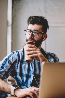 Coworking alanı, uzağa bakarak ve webinar netbook üzerinde bekleyen şaşkın adam masaüstü dizüstü bilgisayar ile oturup sabah kahve içme kulaklık üzerinden yeni çalma listesi zevk düşünceli hipster adam