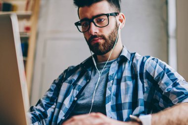 Bilgi için modern teknolojiyi kullanarak ve sesli kitap ciddi hipster adam gözlük netbook üzerinde Internet tarama yolu ile kulaklıklar, dinleme yeni makale arama erkek metin yazarı konsantre