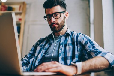 Bilgi için modern teknolojiyi kullanarak ve sesli kitap ciddi hipster adam gözlük netbook üzerinde Internet tarama yolu ile kulaklıklar, dinleme yeni makale arama erkek metin yazarı konsantre
