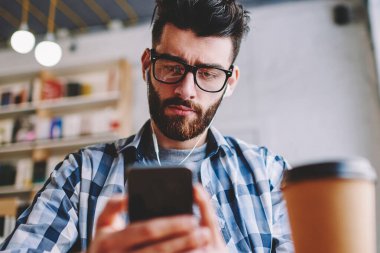 Kahve dükkanında oturmuş ve cep telefonu numarası çevirmeden, gözlük vizyon düzeltme smartphone aşınma Kulaklığımda görüntülü arama yapma hipster adam yoğunlaştı yakışıklı ciddi erkek blogger