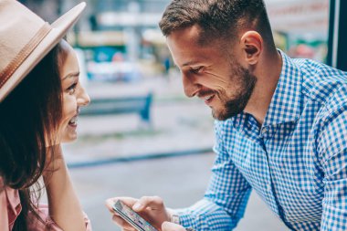 Genç evlilik mutlu bir şekilde gülerek harcama birlikte modern kahve dükkanında serbest zaman. Erkek Smartphone cep telefonu ile sosyal ağlarda profil tarihi kafede neşeli kız arkadaşı ile tadını çıkarırken güncelleştiriliyor