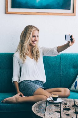 Hippi kız mutlu kadın fotoğraf yolu ile smartphone aygıt ev iç çekmek, sehpa, önünde rahat kanepe otururken telefonla örümcek ağı fotoğraf makinesi ile pozitif selfie fotoğraf yapma gülümseyen