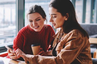 Giyinmiş iki kadın arkadaş toplantı coffee shop smartphone aygıt kullanarak olumlu video blog izlerken zevk, sosyal ağlarda tebliğ hipster kızlar rahat Üniversitesi'nden meslektaşı ile