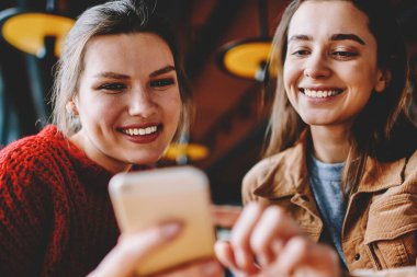 İki gülümseyen kadın blogcular mutlu bin yıllık kadınlar üzerinde boş zaman teknoloji ile dinlenme modern cep telefonu aracılığıyla arkadaşlarınızla sohbet cep telefonu, fotoğraf uygulaması kullanarak yayın oluşturma