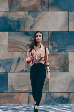 Modaya uygun kıyafet ayakta duvara tanıtım alanı reklam metin mesajları, kamera açık havada seyir casual giyim giymiş genç kadın için çekici hippi kız portresi tam uzunlukta