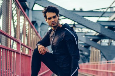 Yakışıklı esmer saç runner dinlenme sırasında kulaklık müzik dinlerken eğitim alarak siyah spor kıyafeti. Mola egzersiz yaptıktan sonra şehirde öğleden sonra genç erkek koşucu