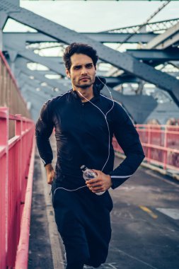 Genç adam şehir Köprüsü'nde şişe su ellerini, kentsel ayarlama ve kulaklık, etkin sağlık ve motivasyon yaşam tarzı ile çalma listesi dinleme çalışan Avrupa atlet ile koşu spor giyim