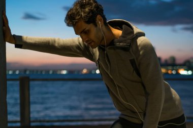 Vücut kas dinleme müzik yolu ile yeni kulaklıklar, Avrupa erkek koşucu dinlenme hızlı şehir sokakları arasında akşam saat sırasında koşu sonra duvarda için kardiyo egzersizleri sonra genç adam duygu yorgun