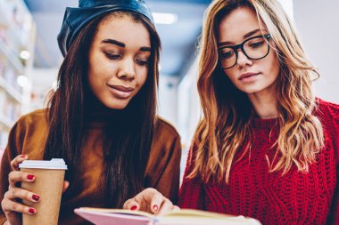 Genç hippi kız ilginç edebiyat birlikte kahve arası kapalı, kafein içecek Eyewear beyaz arkadaş yakınındaki ders kitabı oturuşta bilgi arama ile Afrikalı kadın sırasında okuma.