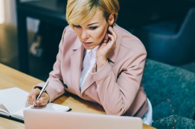 Kadın Olgun girişimci iş webinar online ve Not Defteri'nde aşağı yazma izliyor. Dalgın deneyimli kadın koç ile 4 g laptop çalışırken not defterinde kayıt yapma Internet