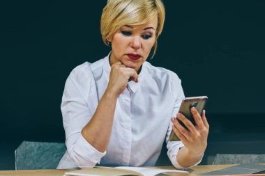Kadın girişimci 50 yaşında smartphone aygıt netbook çalışma video izlerken konsantre. Web sitesi üzerinden 4 g finansal haber okuma ciddi olgun işkadını hücresel internet