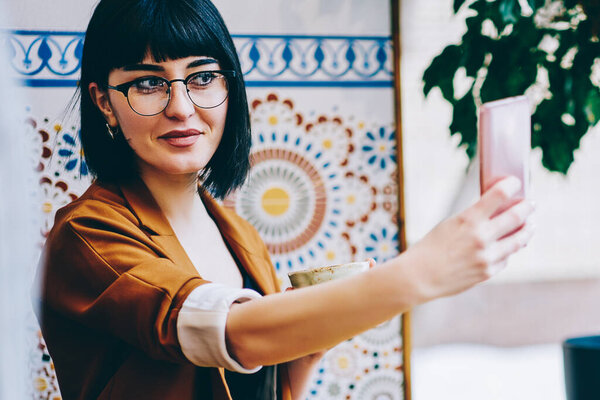 Positive female student posing for selfie photo during lunch at bohemian cafeteria and working on modern netbook, smiling hipster girl making pictures using front camera on smartphone device