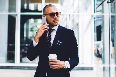 Formal giyim ve şık gözlük duran cam duvar ile bina modern ofisindeki kahve ile banka hesabı kontrol etmek için smartphone: çağrı kendine güvenen genç işadamı giymiş