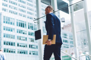 Başarılı iş adamı arkadan görünüm belgeler klasörüyle tutarak ve ofis binası koridorunda gidiş şık takım elbise giymiş. Girişimci formal giyim mali şirket içinde gezinme