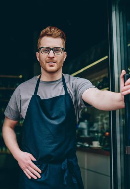 Kameraya gülümseyen ve daha çekici önlük başarılı beyaz barista portresi kafede misafirler. Kamera kendi kafeterya girişinde dururken gülümseyen neşeli erkek garson