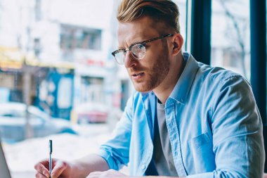 Yetenekli beyaz öğrenci kablosuz internet kullanımı ve kafede oturan notlar iç yapım modern dizüstü bilgisayarda ilginç webinar izliyor. Dalgın genç adam netbook uzaktan çalışma