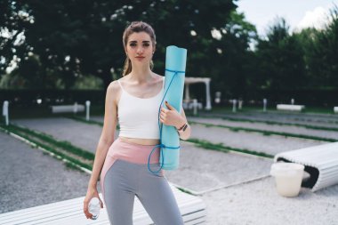 Genç bayan parkta duruyor, yoga seansına hazır. Yoga minderi ve su şişesi tutuyor, şık bir spor kıyafeti giyiyor. Açık havada sakin bir ortam, spor ve refahı ön plana çıkarır.
