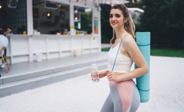 Gülümseyen ve yogaya hazır bir kadın, elinde mavi bir hasırla bir şişe su taşıyor. Renkli süslemelerle dolu bir açık hava kafenin yanında duruyor. Sağlıklı bir hayatı kucaklamaya hazır.