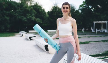 Yoga minderi ve su şişesi tutan genç bir kadın. Gülümsüyor ve yoga seansına hazır. Arka planda banklar ve yeşilliklerle parkta duruyor, dengeli ve sağlıklı yaşam tarzını gösteriyor.