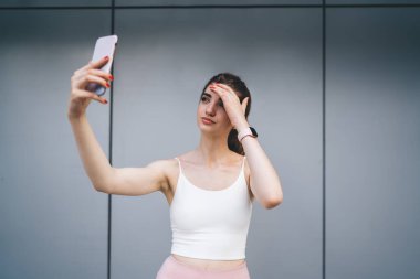 Düşünceli genç beyaz kadın kendini akıllı telefon uygulaması ile fotoğraflıyor. Beyaz spor bluzu ve pembe saati ile saçını düzeltiyor, düşünceli ifadesini yansıtıyor. modern gri arkaplan