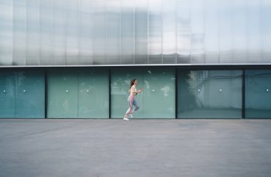 Kulaklıklı genç beyaz kadın şehir ortamında koşuyor, atletik giysiler giyiyor, akıllı telefon tutuyor. Sağlık bilinci ve enerjik yaşam tarzı, camdan pencereleri olan modern bir binanın yanında koşuyor.