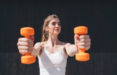 Açık havada egzersiz yapan, turuncu halterlerle yükselen genç beyaz kadın. Beyaz spor sutyen giyiyor, odaklanmış ifade ediyor. Güneşli bir gün, koyu ahşap bir arka plan, yaz sporu