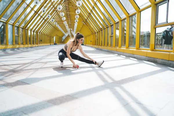 Genç beyaz kadın sabah antrenmanı sırasında şehir ortamında bacak esnetiyor. Spor sutyen ve tayt giymiş, akıllı telefon tutuyor ve kulaklıkla müzik dinliyor, fitness rutinine odaklanıyor.