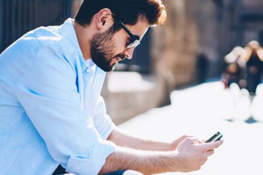 Yan görünüm casual güneş gözlükleri smartphone köknar yayın şehir merkezinde oturan Web sitesinde metin yazarak erkek blogger giymiş. Genç adam 4 Internet bağlantısı üzerinden çevrimiçi arkadaşlarınızla sohbet