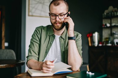 Düşünceli hipster adam sakallı oturma favori kafeterya, erkek öğrenci tarafından ders kitabı içinde café okuma ve düzeltme notu Üniversitesi kampüsünde eğitim ve planları hakkında düşünme gözlük