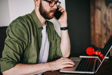 Şık sakallı hippi erkek tarafından smartphone kız arkadaşıyla konuşuyor ve uzay, genç profesyonel programcı adam arayan müşteri site oluşturma hakkında ayrıntılı bilgi tartışmak için coworking içinde dizüstü bilgisayar kullanarak