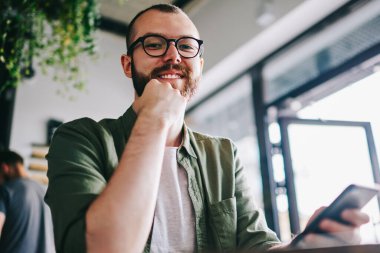 Yakışıklı genç bloger bekleyen arkadaş kafe ve yeni yayın oluşturma, sakallı iken cep telefonu elinde tutan ve sohbet hipster adam kamera seyir gözlük gülümseyen portresi