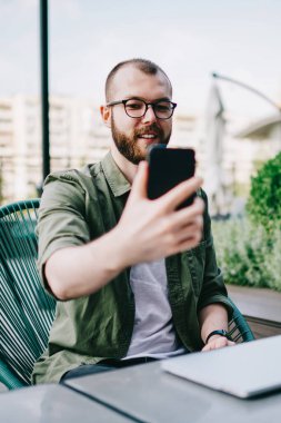 Sokak kafe masada oturan ve yeni blog için video yapmak için smartphone kullanarak hipster adam gülümseyen hareket eden telefon tutarak ve modern ön kamera ile selfie fotoğraf çekmek yakışıklı sakallı erkek