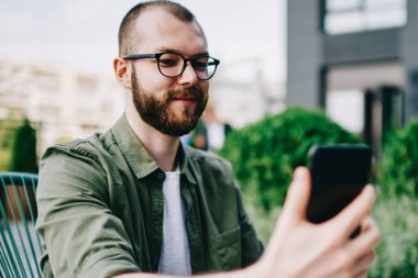 Trendy erkek holding cep telefonu cep telefonu kamera ve kendini açık havada kafede oturan ve selfie fotoğrafı tıklatın smartphone kullanarak beyaz hipster adam alarak fotoğraf gülümseyen elinde sakallı