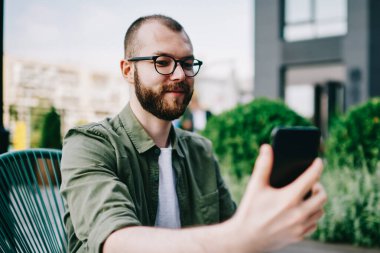 Trendy erkek holding cep telefonu cep telefonu kamera ve kendini açık havada kafede oturan ve selfie fotoğrafı tıklatın smartphone kullanarak beyaz hipster adam alarak fotoğraf gülümseyen elinde sakallı