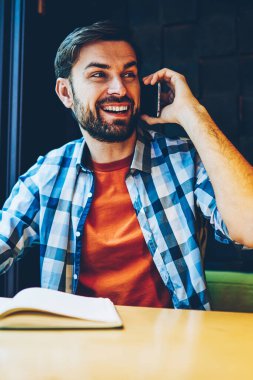 Pozitif sakallı hippi adam smartphone aygıt üzerindeki telefon görüşmesi sırasında gülüyor. Uzay coworking oturan telefonda konuşurken Not Defteri'nde bilgi yazma rahat kıyafet içinde neşeli erkek blogger