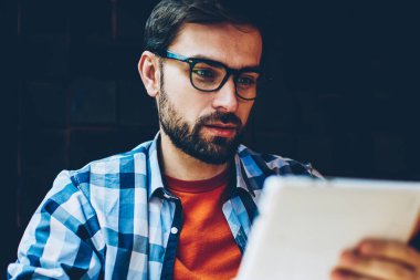 Dalgın sakallı genç adam 4 g üzerinden Web sitesinde haber okumak için dijital tablet Internet bağlı. Erkek blogger gözlük vizyon düzeltme Dokunmatik ped cihaz yayınında İndiriyor yoğunlaştı