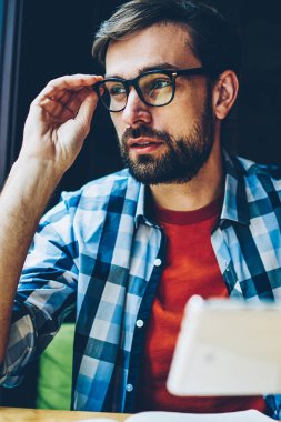 Uzağa bakıyor ve kendi blog geliştirmek için fikirleri üzerinde düşünme dokunmatik yüzey ile düşünceli sakallı genç adam. Gözlük vizyon düzeltme modern tablet elinde tutan durulması hipster adam