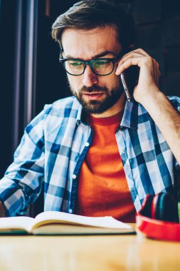 Kızgın genç adam kitap alan coworking içinde vasıl okul sırası oturan smartphone aygıt üzerinde iletişim gözlük sakallı. Telefon görüşmesi sırasında konuşurken rahat kıyafet rahatsız hipster adam giymiş