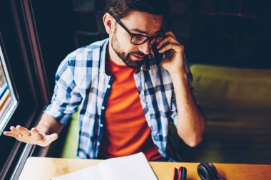 Üstten görünüm sakallı genç adamın smartphone vasıl okul sırası yer coworking yılında ders kitabı ile oturup telefon görüşmesi sırasında konuşurken rahat şık gömlek giymiş. Hipster adam cep üzerinde arama