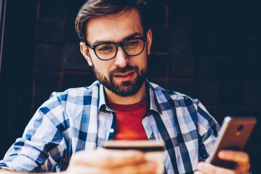 Dalgın casual giyinen hipster adam web sitesinde sipariş yapmak için kartvizit smartphone telefon numarasını çevirme. Yapım para hareket online üstünde hareket eden telefon içinde coworking oturan genç adam