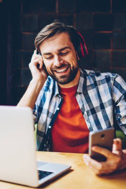 Mutlu sakallı genç modern kulaklık smartphone aygıtında indirilen çalma listesinden müzik dinleme adam. Dijital dizüstü bilgisayar başında oturan radyo web sitesinde ses şarkı zevk neşeli erkek blogger