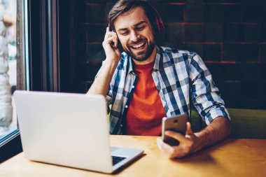 Pozitif sakallı genç adam favori ses çalma listesinden şarkıyı modern Kulaklığımda Smartphone'da indirilebilir. Mutlu hipster adam dinleme müzik radyo oyuncu laptop oturan telefonu