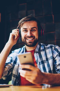 Mutlu sakallı erkek blogger smartphone için Bluetooth kulaklık uygulamada müzik dinleme şarkıları bağlı iken kameraya gülümseyen portresi. Neşeli hipster adam zevk çalma listesi