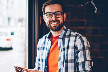 Profil yolu ile 4 g Smartphone cep telefonu ile güncelleştirilirken kameraya gülümseyen gözlük içinde başarılı sakallı hippi blogger portresi Internet. Mutlu genç adam mobil telefon elinde tutarak rahat şık gömlek