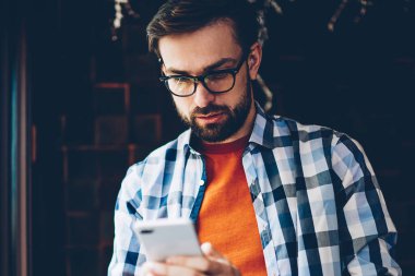 Dalgın sakallı hippi blogger gelen bildirim okuma ve ağlar Smartphone cep telefonu ile sohbet bağlı 4 g için Internet. Genç adam cep telefonu online video izlerken rahat gömlek giymiş