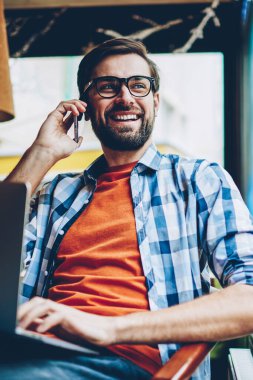 Smartphone üzerinde arama ve pencereden dışarı bakıyor telefon görüşme sırasında gülüyor neşeli sakallı hippi adam. Netbook çalışırken arkadaşıyla telefonda konuşurken olumlu erkek blogger