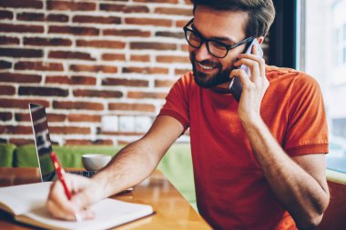 Neşeli smartphone aygıt üzerinde arama ve metin bilgileri dizüstü cihaz alan coworking içinde oturan Not Defteri'nde yazma genç adam sakallı. Telefonla konuşurken ve notlar olumlu hipster adam