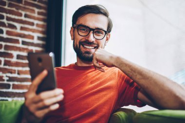 Başarılı sakallı erkek blogger smartphone aygıt ücretsiz 4 g internet bağlantısı üzerinden app karşıdan yüklerken kameraya gülme portresi. Pozitif hipster adam cep üzerinde online ödeme yapma