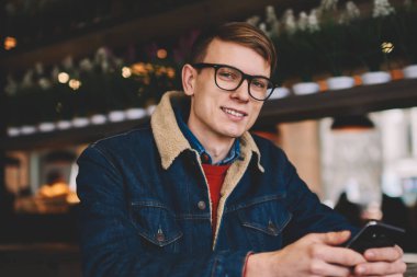 Yarım uzunluk portre denim ceket şık kahve dükkanında dinlenme 4g internet bağlantısı ile Smartphone cep telefonu ile sosyal ağlarda profil güncelleştirilirken kameraya gülümseyen başarılı erkek Blogger